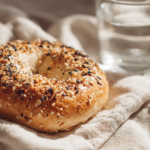 Close-up of a golden brown protein bagel with everything seasoning on a white napkin, alongside a glass of water, bright natural light.