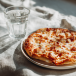 A close-up shot of a freshly baked keto cottage cheese pizza crust, topped with melted cheese and fresh basil, on a white plate with a neutral linen napkin and a glass of water.