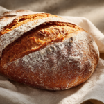 Freshly baked sourdough loaf with a clean central slash scoring dusted with flour, on white cloth with water glass nearby.